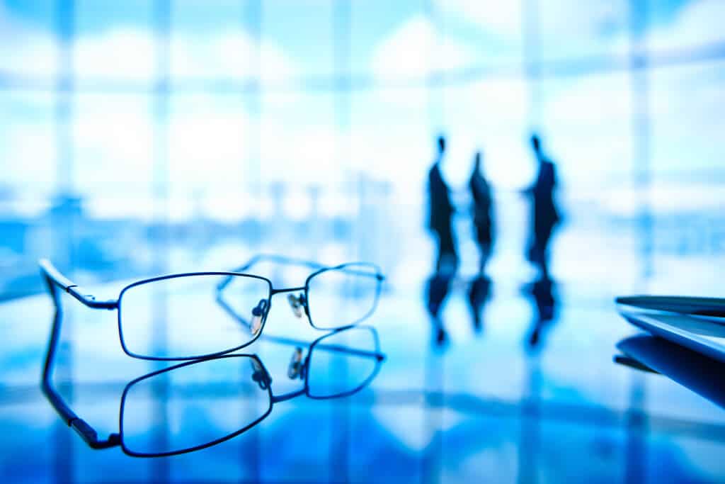 Óculos sobre mesa com silhuetas ao fundo em ambiente corporativo