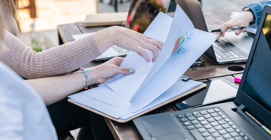 Pessoas em uma mesa de trabalho manuseiam documentos com gráficos, cercadas por notebooks e celular.