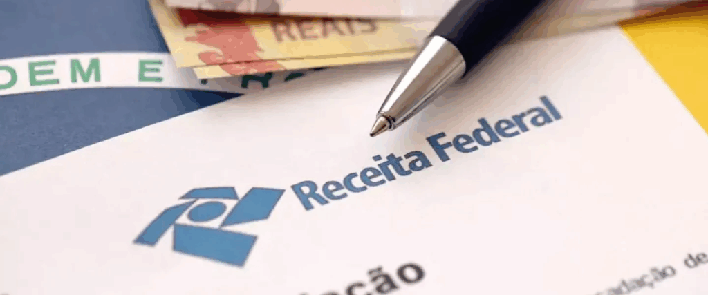 Close de um documento da Receita Federal com uma caneta sobre a folha e notas de real ao fundo.