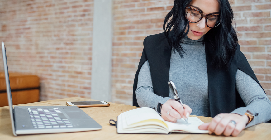 Mulher sentada à mesa escreve em um caderno aberto, com notebook e celular ao lado, em ambiente de trabalho com parede de tijolos aparentes.