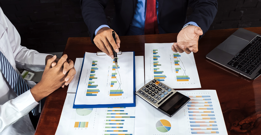Profissionais reunidos em uma mesa analisam gráficos e relatórios financeiros impressos, com calculadora e notebook ao redor.