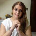 Mulher de cabelos castanhos com as mãos unidas, usando uma blusa branca, pulseira dourada e relógio de pulso rosa.