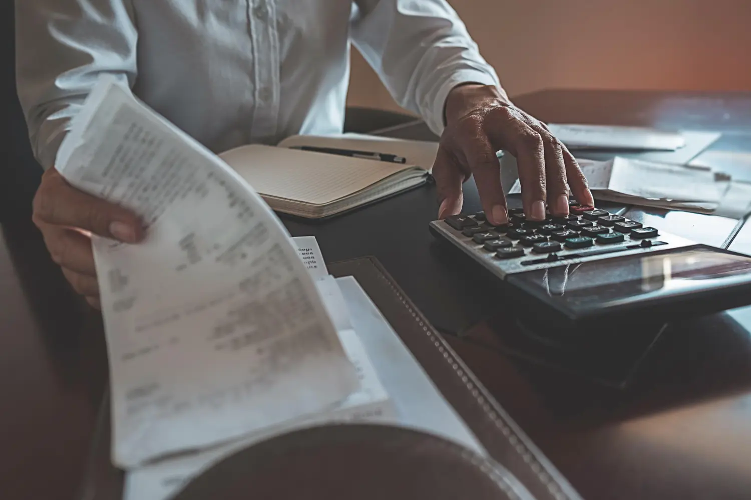 Mão de homem em camisa branca operando uma calculadora com uma folha de papel na outra mão.