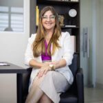 Retrato profissional de mulher sorrindo sentada em escritório corporativo, utilizando óculos e traje social