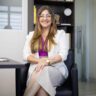 Retrato profissional de mulher sorrindo sentada em escritório corporativo, utilizando óculos e traje social