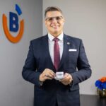 Homem de meia idade, de óculos, terno azul e gravata vinho, segurando xícara de café com o logo da bandeira.