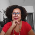 Mulher de meia-idade, morena, óculos, cabelos cacheados, blusa vermelha, colar e brincos.