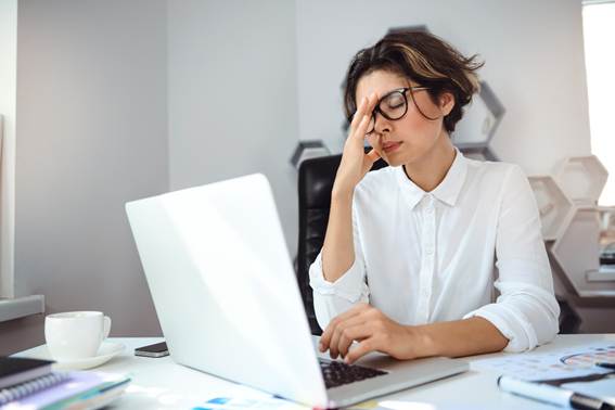 Mulher de óculos e camisa branca sentada em um escritório de frente para um notebook aberto. Ela está com a mão no rosto e os olhos fechados, expressando forte cansaço, estresse ou frustração com o trabalho.