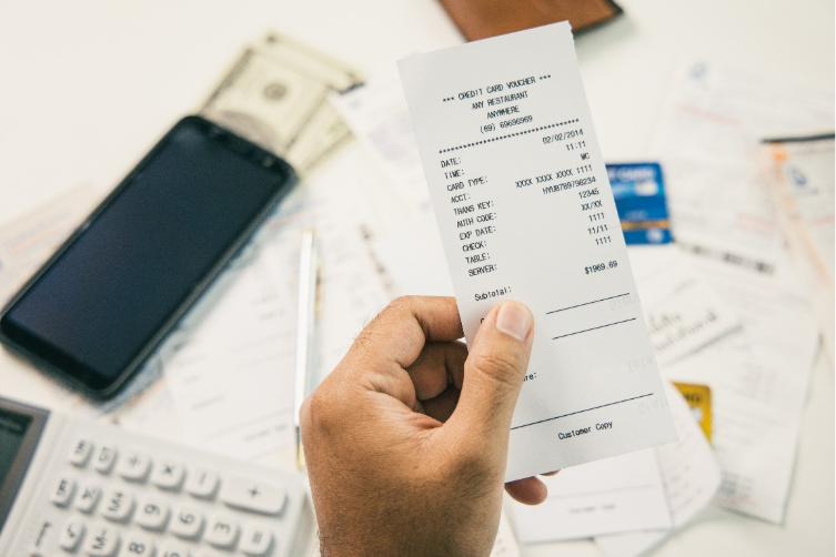 Mão segurando cupom fiscal com itens de compra, celular, dinheiro e calculadora ao fundo.