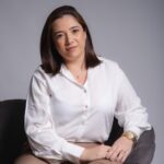 Retrato profissional de mulher sorrindo sentada em escritório corporativo, utilizando óculos e traje social