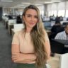 Retrato profissional de mulher sorrindo sentada em escritório corporativo, utilizando óculos e traje social