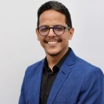 Retrato de um profissional sorridente usando óculos, camisa preta e blazer azul, em fundo neutro, representando liderança, consultoria e atuação na área contábil e tributária.