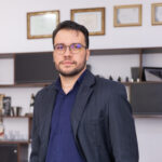 Homem em pé usando blazer azul-escuro, camisa azul-marinho e calça cinza, com uma das mãos no bolso, posando em um escritório moderno. Ao fundo, há prateleiras, quadros com certificados e mobiliário corporativo desfocados.