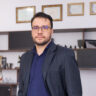Homem em pé usando blazer azul-escuro, camisa azul-marinho e calça cinza, com uma das mãos no bolso, posando em um escritório moderno. Ao fundo, há prateleiras, quadros com certificados e mobiliário corporativo desfocados.