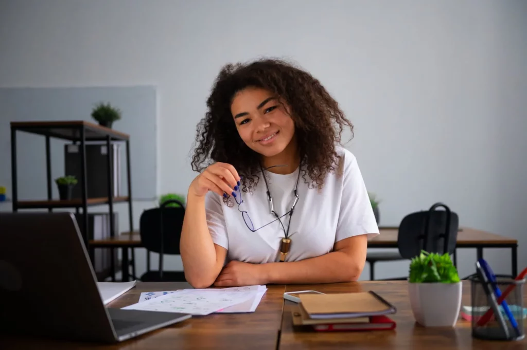 Mulher sorrindo sentada à mesa de trabalho, com notebook, documentos e óculos nas mãos, em ambiente de escritório.