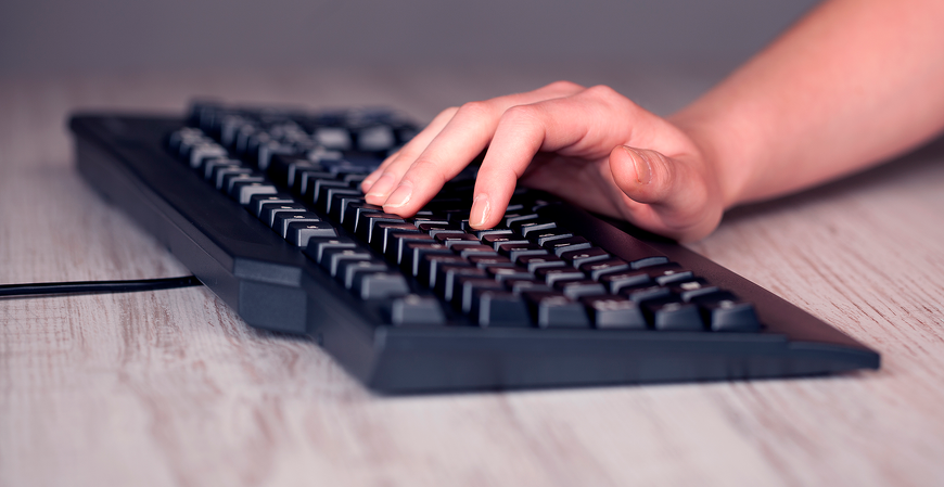 Close-up de uma mão digitando em um teclado de computador sobre uma mesa de madeira.