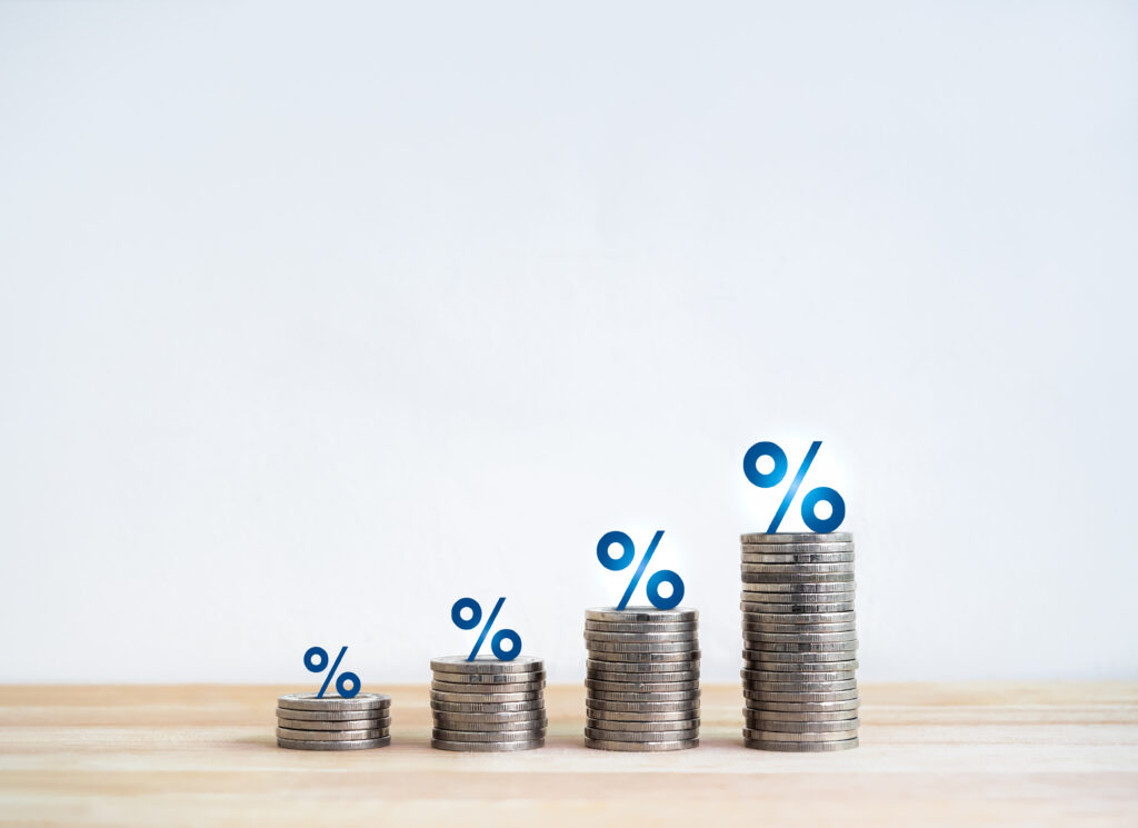Quatro pilhas de moedas prateadas em ordem crescente de altura sobre uma superfície de madeira clara, cada uma com um ícone azul de porcentagem (%) posicionado no topo, simbolizando crescimento de taxas ou percentuais.