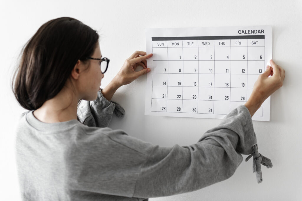 Vista de costas de uma mulher de cabelos castanhos, óculos e moletom cinza, fixando um calendário de papel em uma parede branca com as duas mãos.